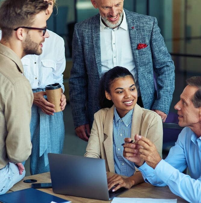 Various individuals gathered around a table and laptop chatting